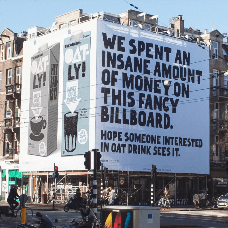 A giant Oatly billboard on a building with the photo of the product with the caption: "We spent an insane amount of money on this fancy billboard. Hope someone interested in oat drink sees it."